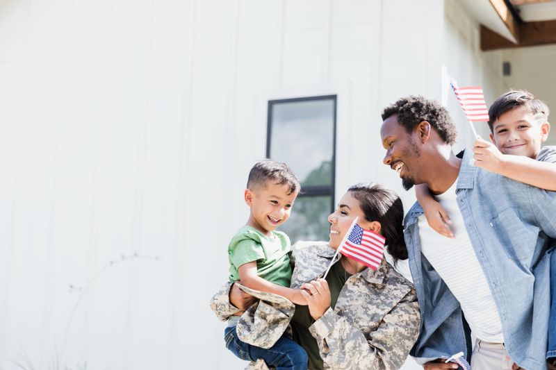 A female soldier holds her young son who has greeted her upon her return from army assignment. Her husband and other son smile cheerfully.