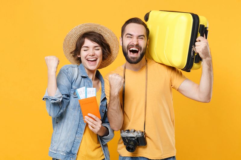 Happy young tourists couple friends guy girl isolated on yellow background. Passenger traveling abroad on weekends. Air flight journey concept. Hold passport tickets suitcase doing winner gesture