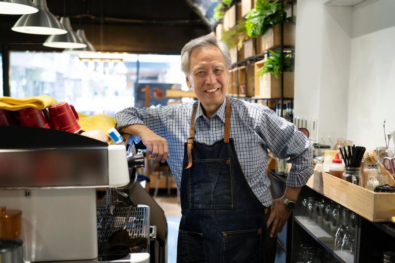 An older Asian barister is leaning his arm on the coffee machines, behind the counter of a cafeteria wearing a smart blue apron