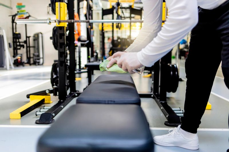 Young man cleaning and disinfecting with sanitizer the gym equipment. Hands of men at the modern gym using the cloth and disinfection liquid in spray bottle to clean the exercising machines in the gym