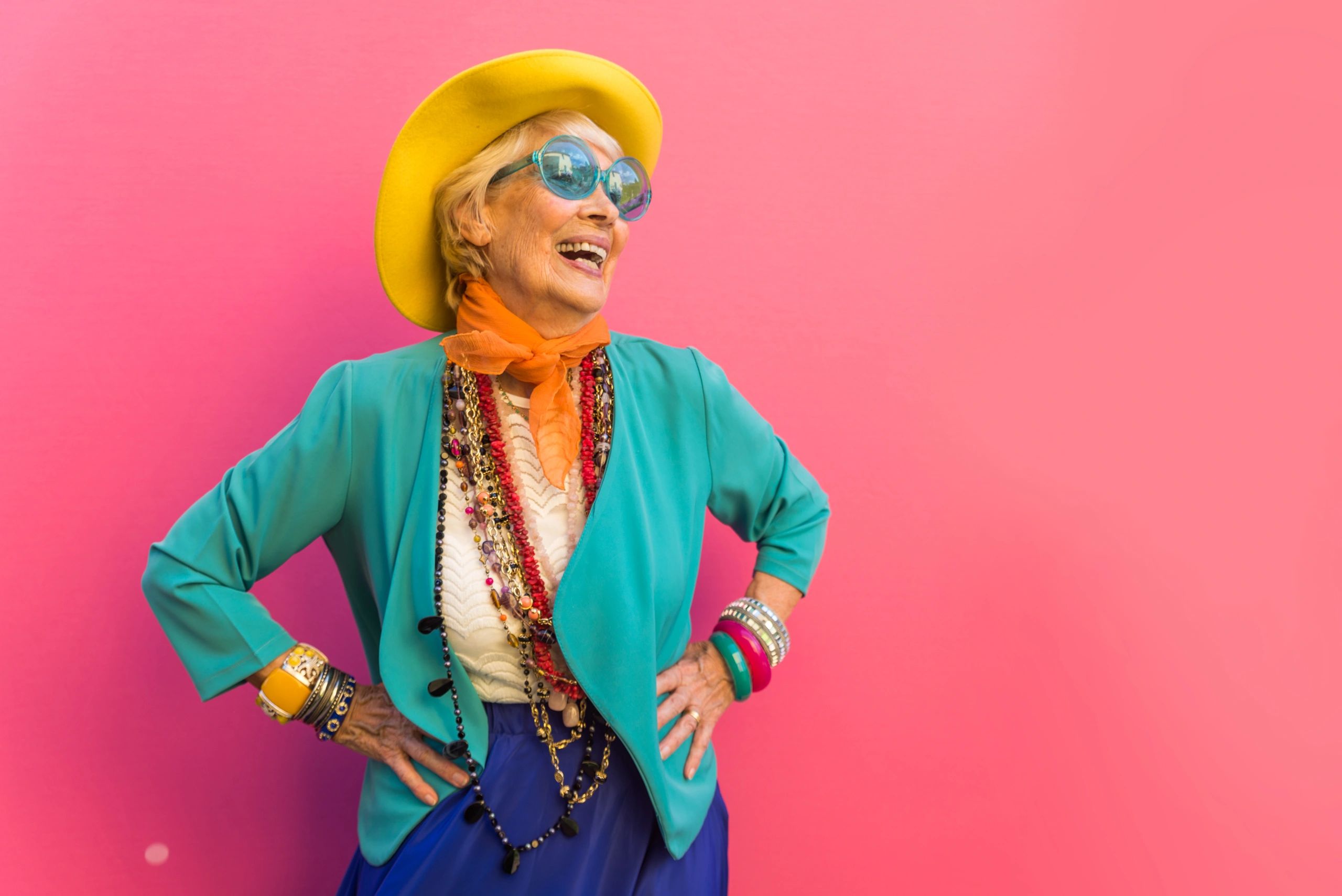 Elderly woman joyfully dressed in vibrant, colorful clothes and accessories against a pink background.