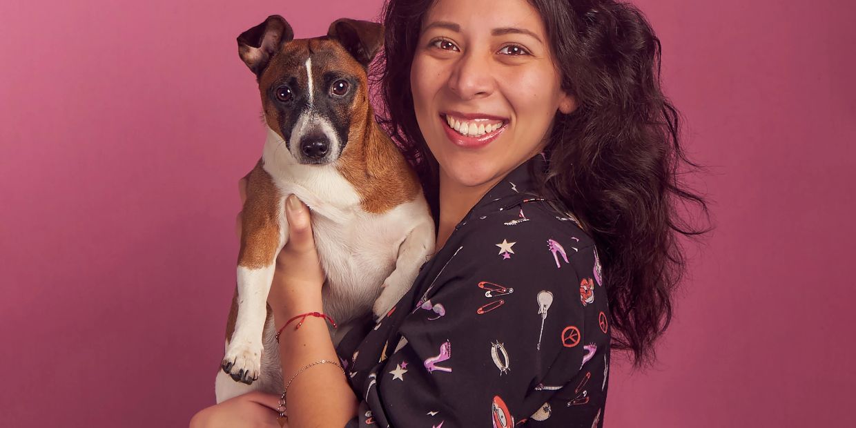 Woman smiling, holding a dog against a pink background.