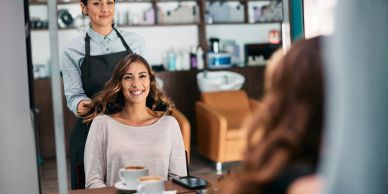 A woman smiles at her reflection while a hairstylist stands behind her.