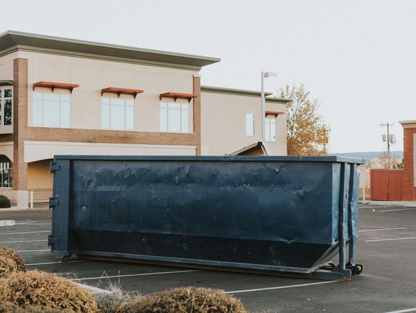 A large blue dumpster sits empty in a quiet parking lot near buildings.