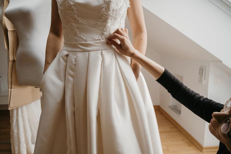 Young bride standing in bridal studio and trying wedding dress with her tailor