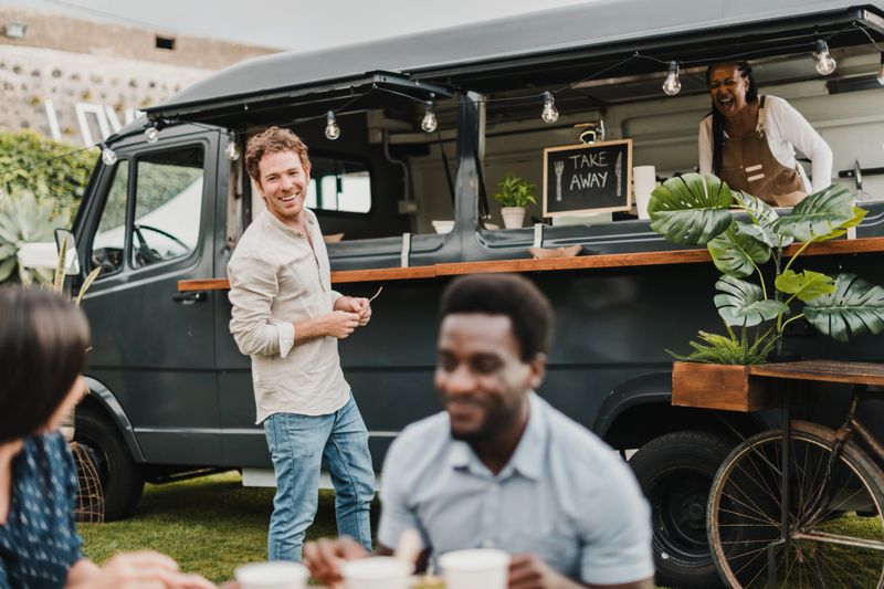 Happy male customer smiling and speaking with diverse friends while standing near food truck and making order to laughing black woman in park