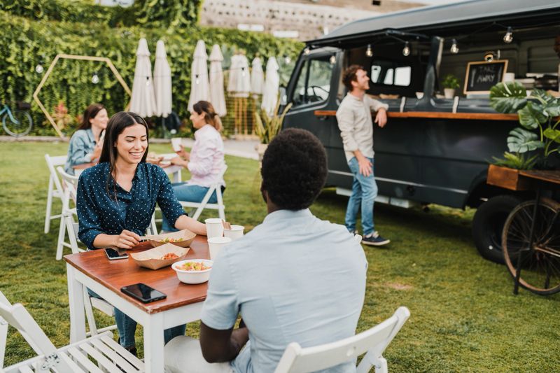 Cheerful woman smiling while sitting at table and having lunch with black boyfriend on summer weekend day near food truck in park