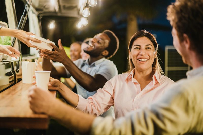 Happy Hispanic female with takeaway drink smiling and talking with male friend while resting outside food truck in evening