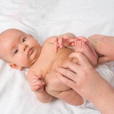 A baby lying on a white blanket participates in gentle infant physiotherapy exercises to promote tension balance and gross motor skills. 