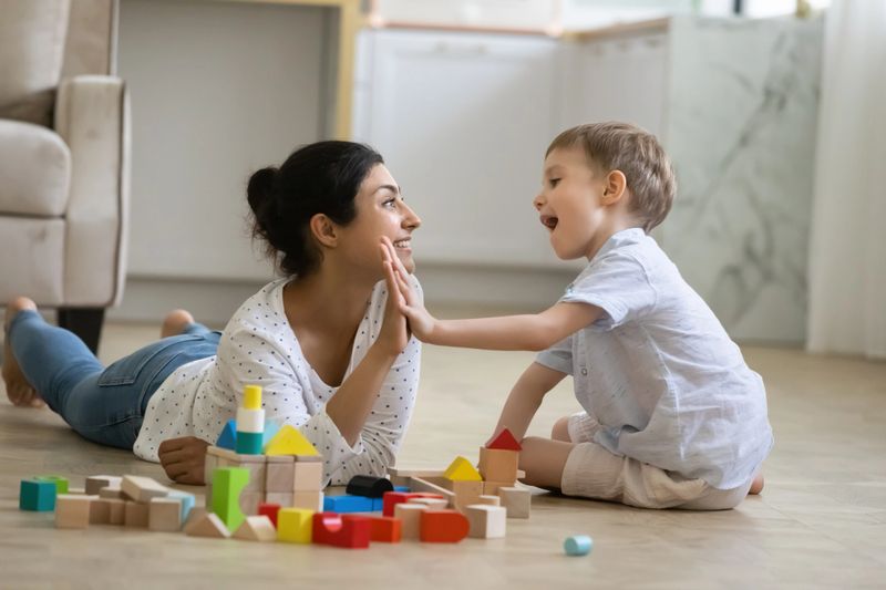 Happy nanny giving high five praise to excited preschool kid boy for completing toy tower on warm floor. Indian babysitter and kid playing at home, constructing building from small wooden blocks