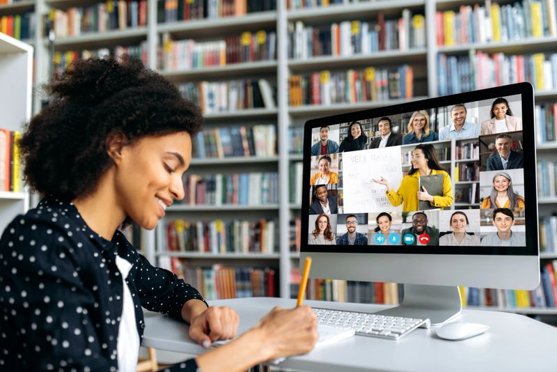A mixed-race student of color watching an educational video call with a diverse group of online learners on screen.