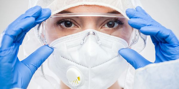 A woman is wearing PPE. A white respirator, blue exam gloves, a white bonnet, and goggles.