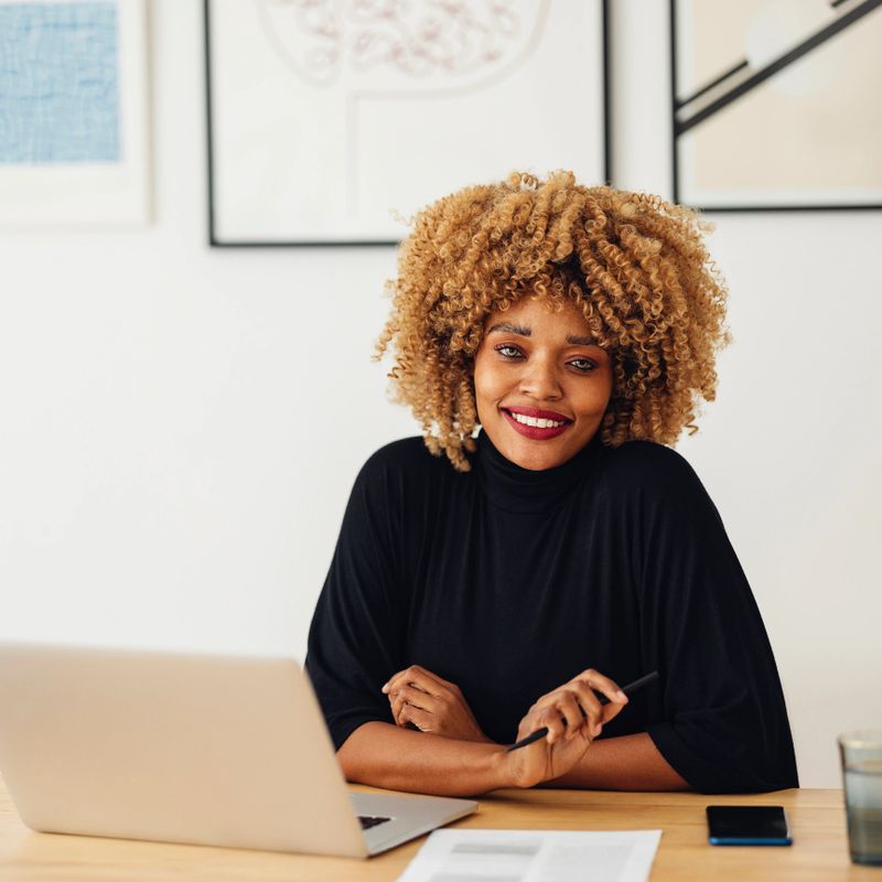 Happy Afro-American woman in a black turtleneck is sitting in a home office and looking at the camera. It's morning or later in the day. She might be a designer or an architect. The room has a modern interior and the wall is full of paintings. The woman might also be a student who is e-learning a course. She is holding a pen. There is a laptop in front of her.