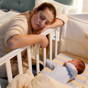Tired mother resting on crib railing, watching her baby sleep peacefully.