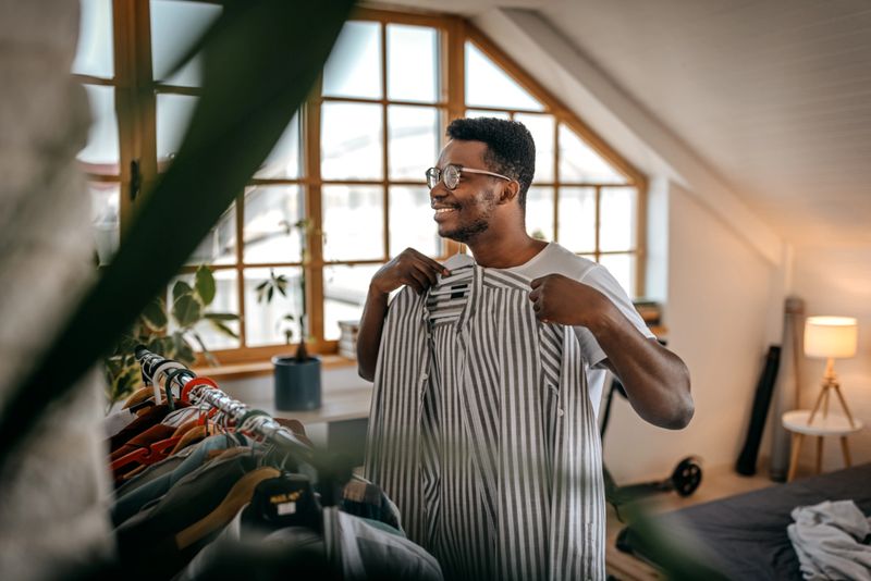 Young businessman selecting and looking at clothes on hangers at home and choosing what to wear by checking on self