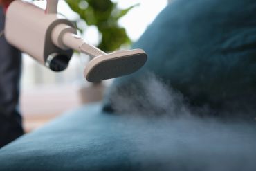 Steam cleaner sanitizing a blue fabric sofa with visible steam.