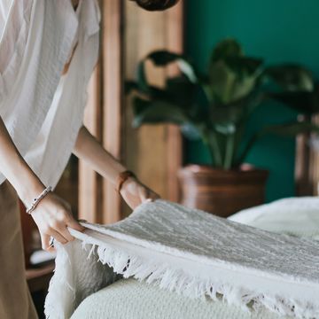 A person making a bed with a white fringed blanket in a cozy room.