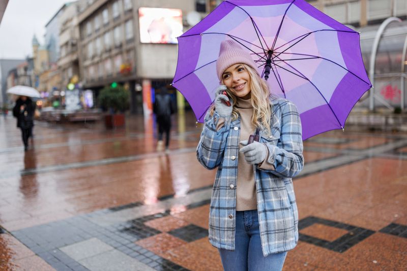 Beautiful young woman walking through the city in the rain, carrying umbrella and talking on the phone