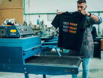 Man holding a black t-shirt with colorful test prints in a printing workshop.