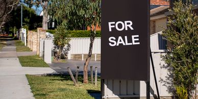 Black 'For Sale' sign outside a suburban house on a sunny day.