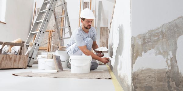 A construction worker plastering a wall indoors wearing a white hard hat.