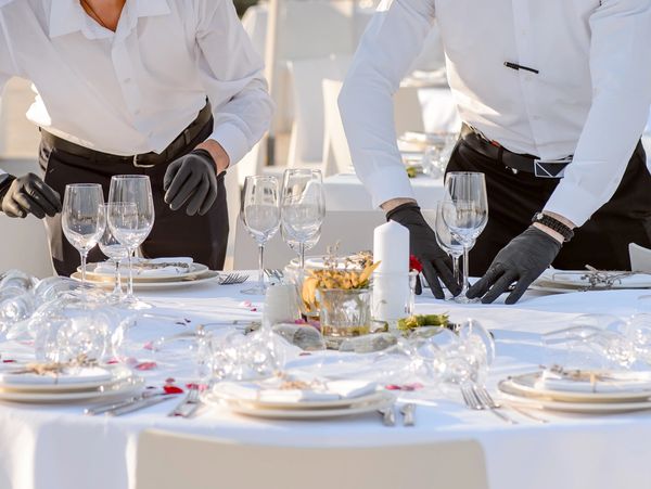 Waiters in black gloves set up an elegant outdoor table with glassware and plates.