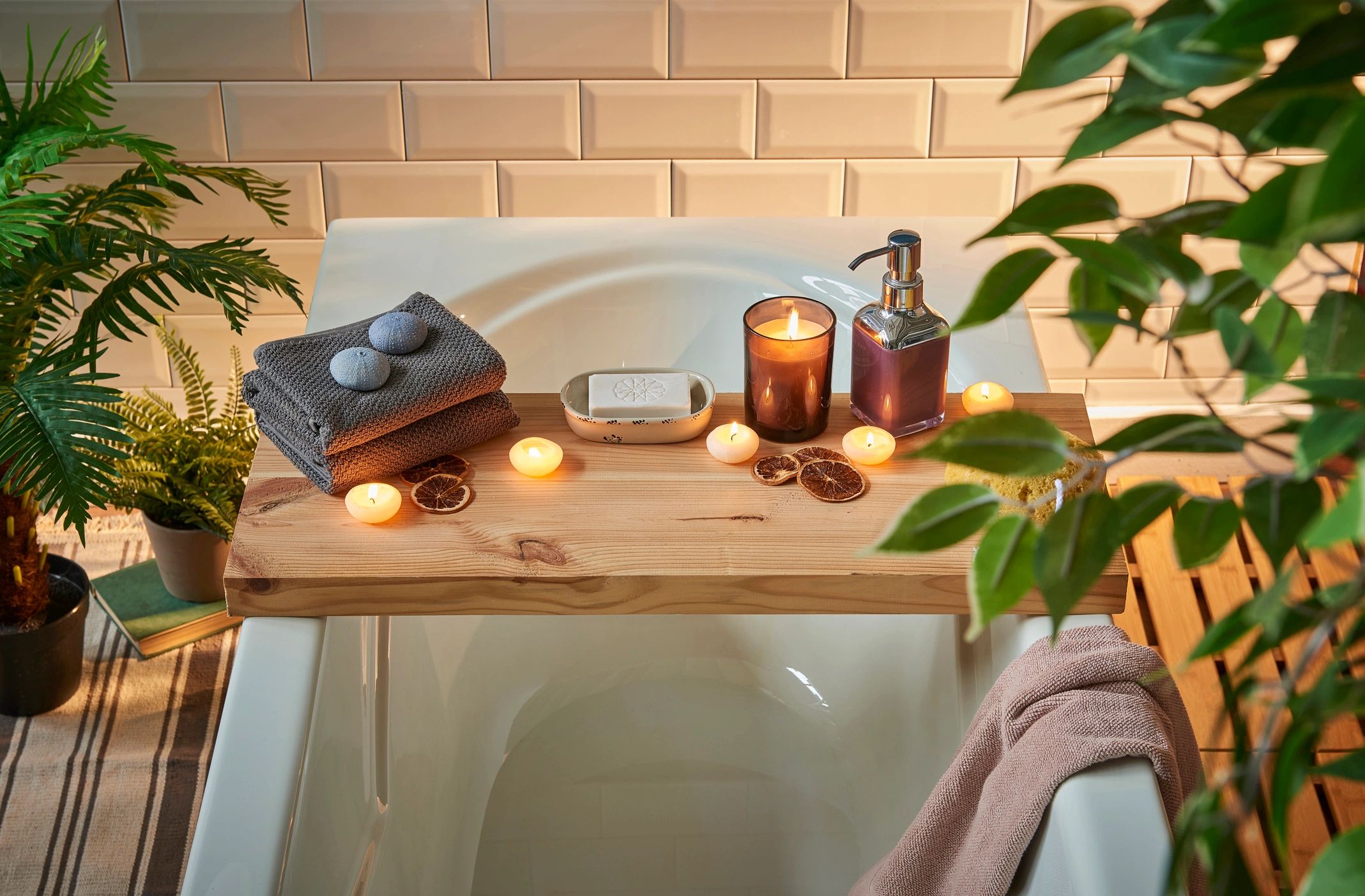 Bathtub surrounded by plants, a wooden tray laying across. There are lit candles and bath products. 