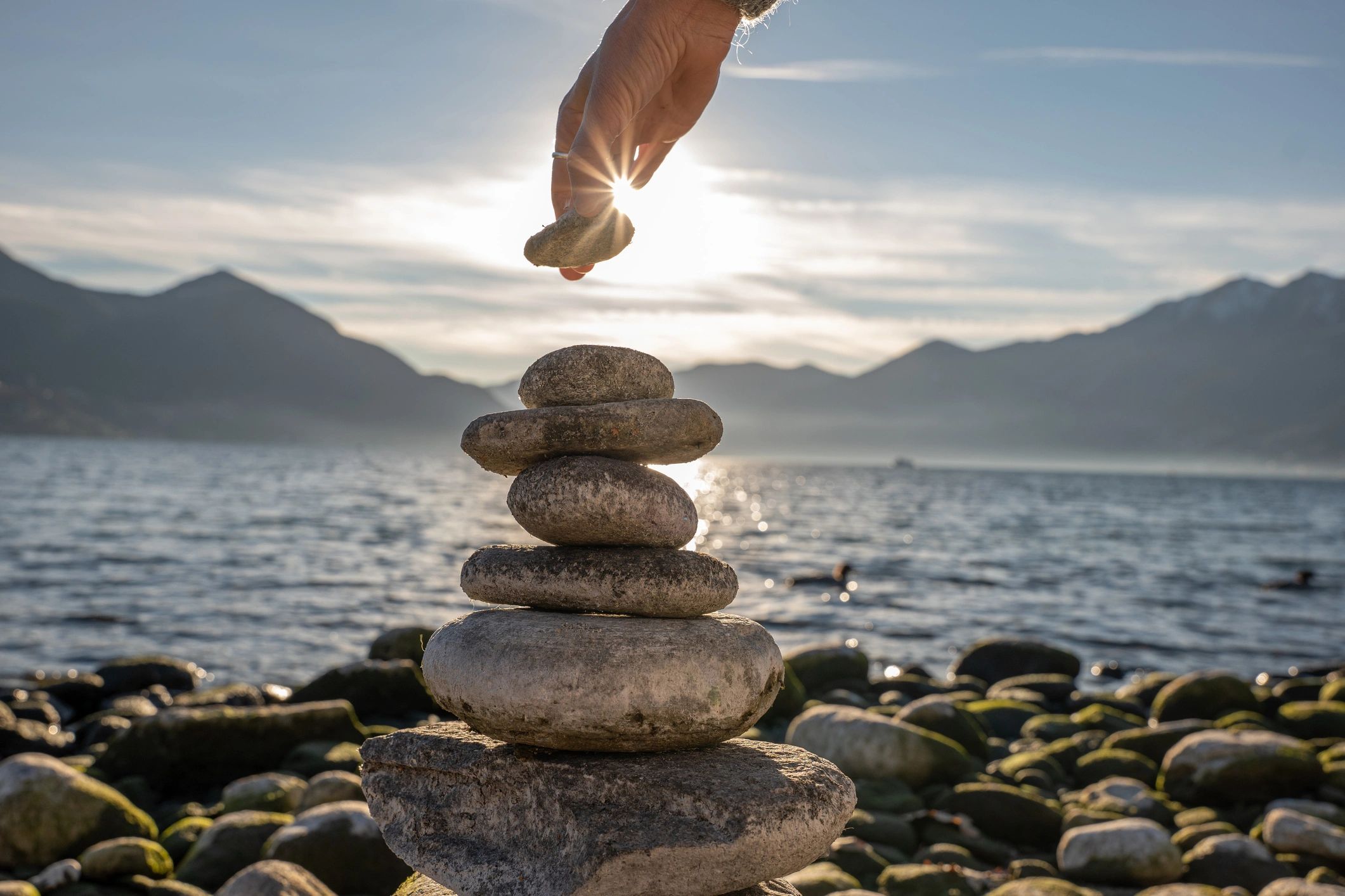 Hand stacking stones by the water with mountains in the background.