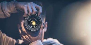 Person holding a camera lens close to the face in a dimly lit setting.