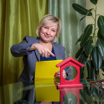 Smiling woman pointing at a yellow folder with a red house model on the table.