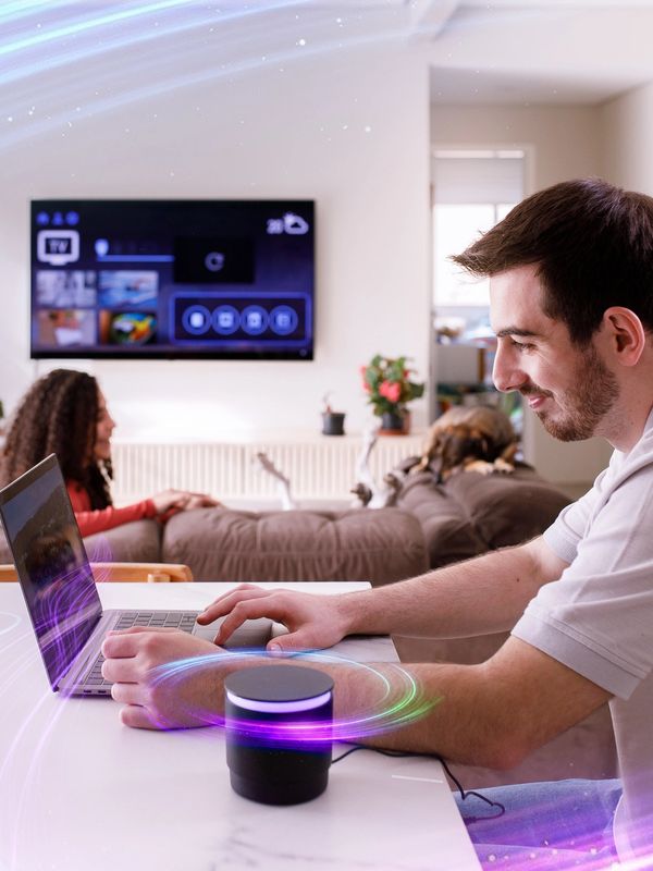 Man using laptop and smart speaker with woman relaxing in background.