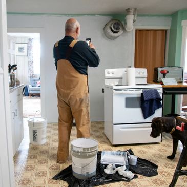Man in overalls taking a photo while painting a kitchen wall with a dog nearby.