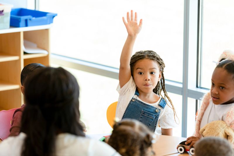 A cute little girl raises her hand to ask or answer a question while sitting at the table with other students and a teacher.