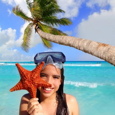 Smiling girl with snorkeling mask holds a red starfish by the beach under a palm tree.