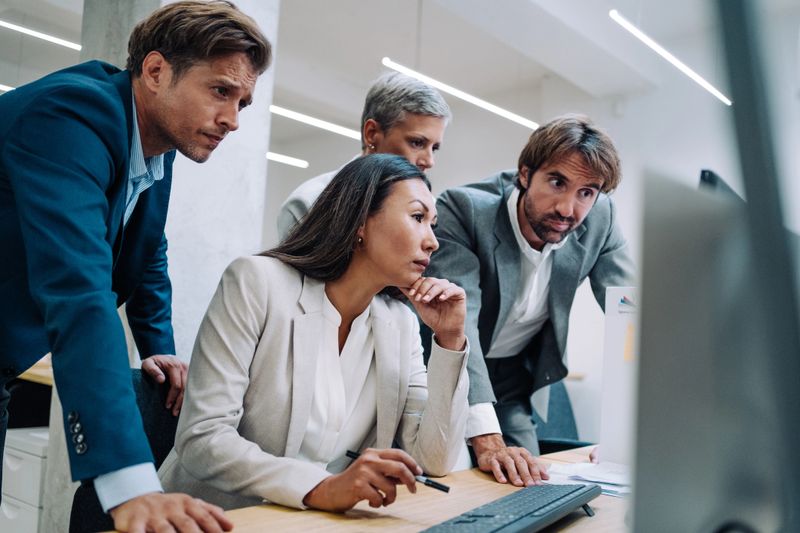 Shot of a group of colleagues using computer together at work. Four coworkers working on a computer in the office. Multi-ethnic corporate team using computer in modern office. Serious business people cooperating while working on PC.
