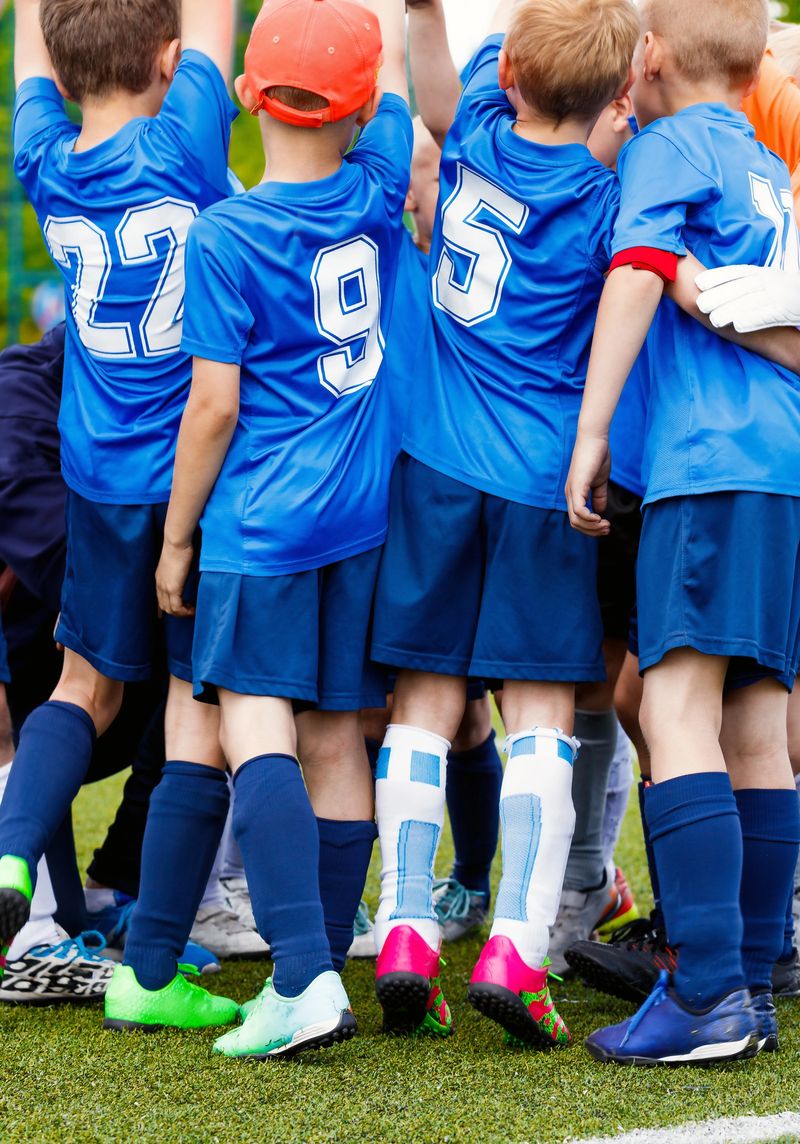 Vertical picture of happy boys in sports team celebrating success together. School kids in blue sporty jersey shirts with white numbers. Children playing sports on grass venue