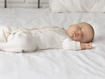 A baby peacefully sleeping on a white bed in a white outfit.