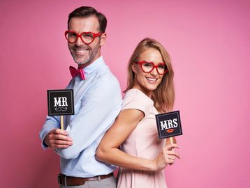Happy couple wearing heart-shaped glasses hold Mr and Mrs signs against pink background.