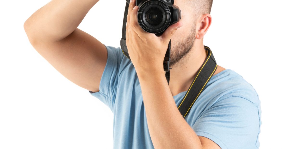 Man in blue shirt taking photo with DSLR camera against white background.