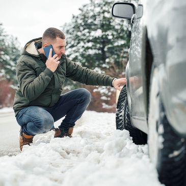 Man checking car tire in snowy conditions while talking on phone.