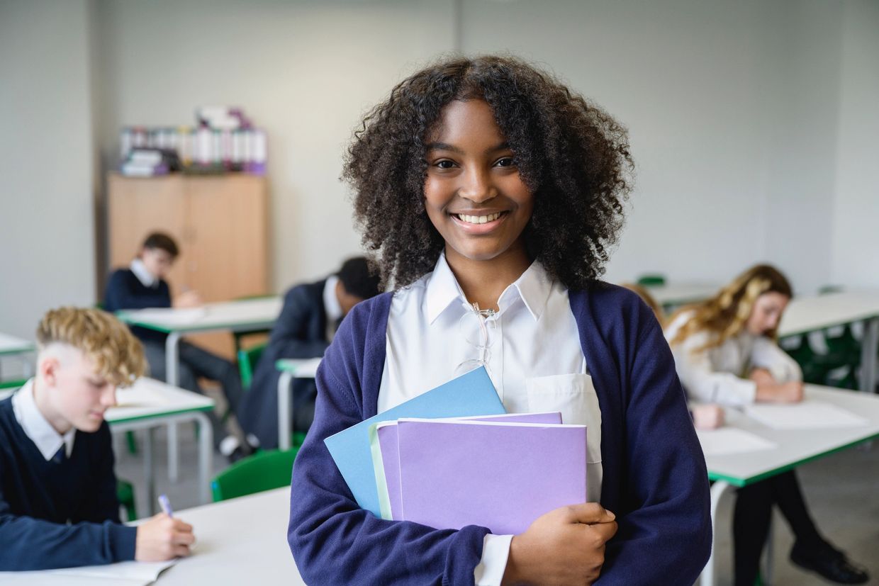 Smiling student holding folders in classroom with peers studying in background.