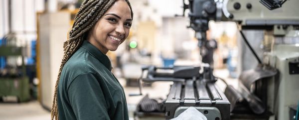 A smiling woman with braids works in a machine shop.