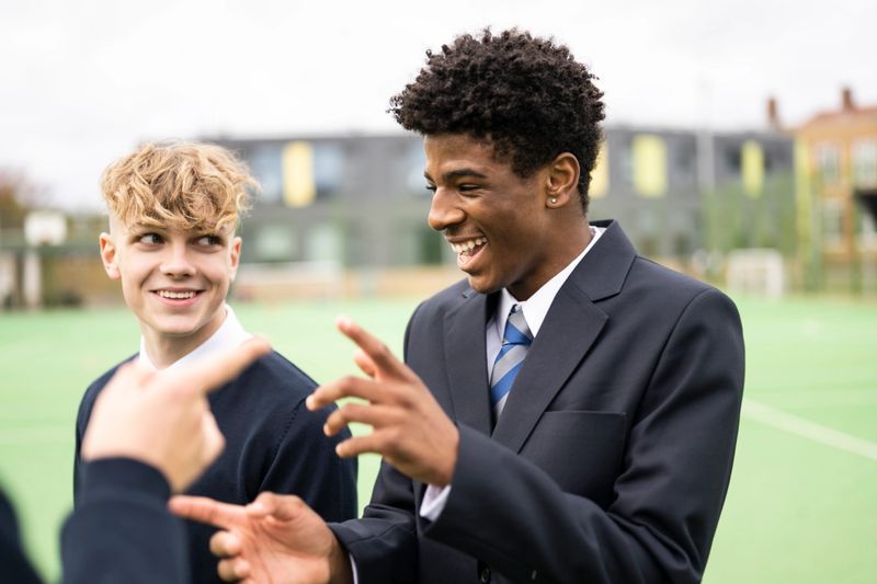 Close-up of teenage students in uniforms standing outdoors on sports field talking, laughing, and gesturing.