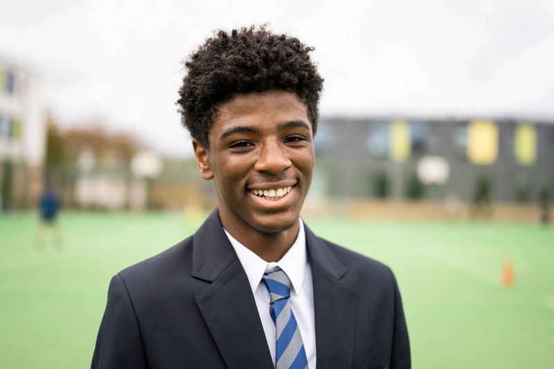 Head and shoulder front view of 15 year old student in shirt, necktie, and navy blue blazer standing outdoors on sports field and smiling at camera.