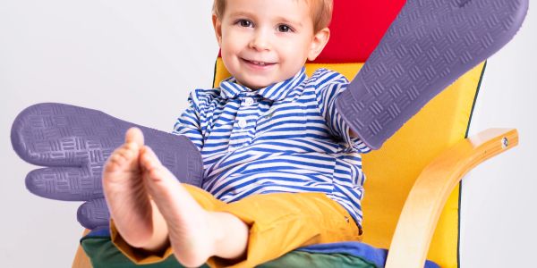 Smiling child sitting on a colorful chair wearing oversized purple mittens.