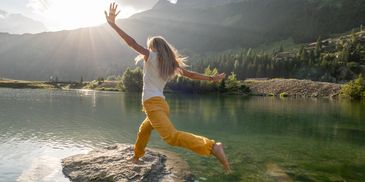 A woman joyfully jumps from a rock into a scenic mountain lake at sunset.