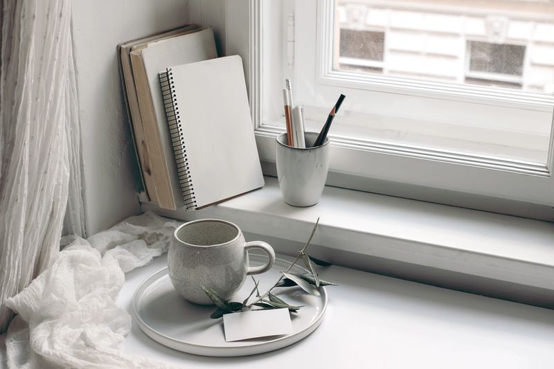 Home office still life. Blank diary and business card mockup. Cup of coffee, olive tree branch on tray. Books and pencils in ceramic mug on window sill. Scandinavian interior, cotton muslin curtain.