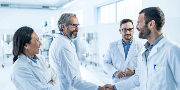 Four scientists in lab coats shaking hands and smiling in a laboratory.