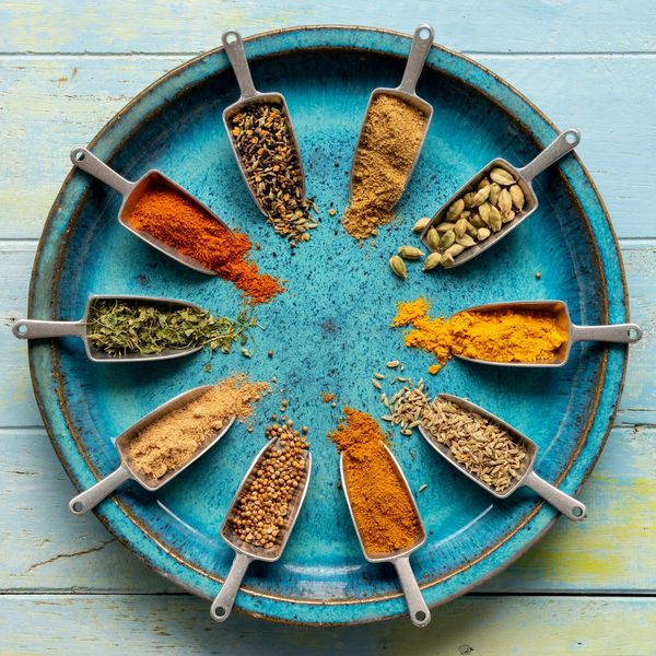 Metal scoops with various colorful spices arranged in a circle on a blue plate.