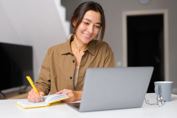 
The image shows a smiling young woman, with dark hair pulled back, sitting at a table with a laptop